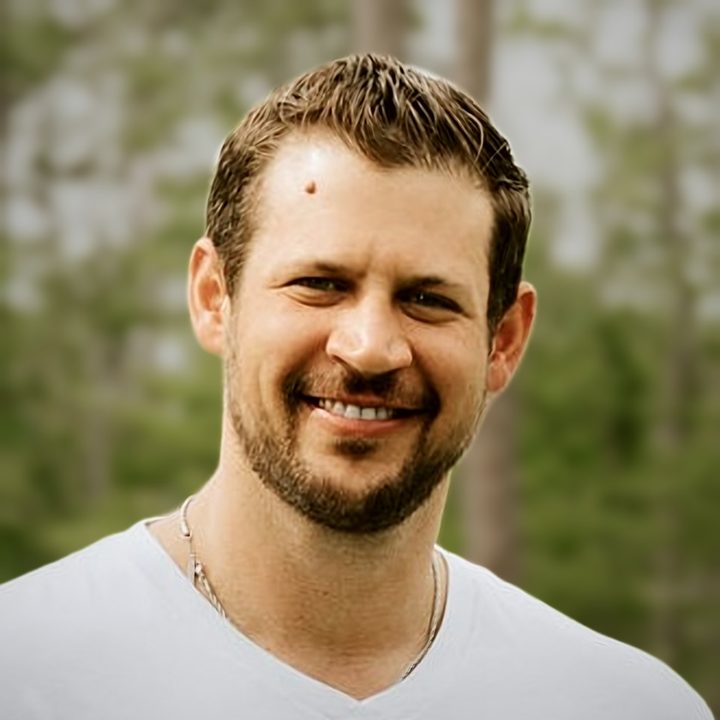 Headshot of Jeremy Van Winkle smiling outdoors in a white V-neck T-shirt, with blurred green trees behind him
