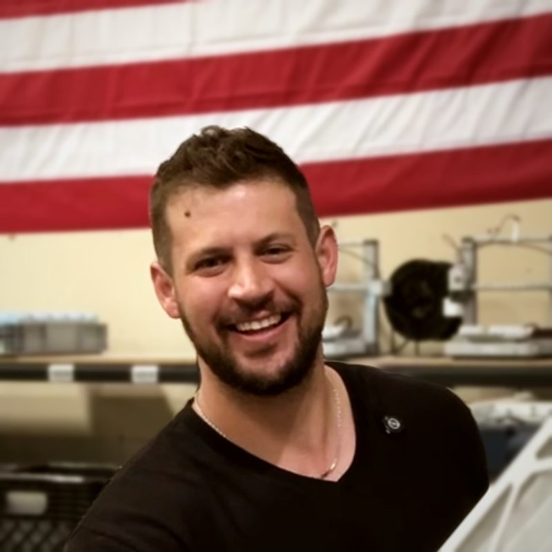 Jeremy Van Winkle smiling in a black T-shirt in a workshop, with a large American flag on the wall and equipment on shelves behind him
